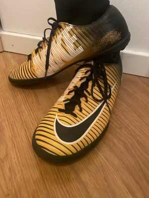 Nike Mercurial gula futsal skor - Säljer ett par Nike Mercurial fotbollsskor i gult och svart med vita detaljer och klassisk Swoosh-logga. Skorna har snörning och en smidig, låg profil som passar perfekt för snabba rörelser på planen. Snyggt randigt mönster och sportig design.