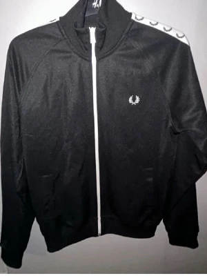 Svart track jacket från Fred Perry - Snygg svart track jacket från Fred Perry med vit dragkedja och logga på bröstet. Axelpartiet har vita band med Fred Perry-kransar längs hela ärmen. Jackan har hög krage och ribbade muddar. Perfekt för en sportig och trendig look.