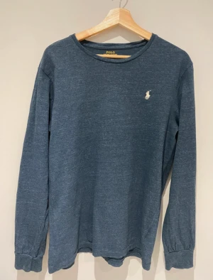 Mörkblå långärmad tröja från Polo Ralph Lauren - Mörkblå långärmad tröja från Polo Ralph Lauren med klassisk vit broderad logga på bröstet. Tröjan har rund halsringning och är tillverkad i mjukt material som passar perfekt till en avslappnad stil.