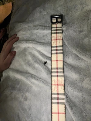 Rutigt bälte med metallspänne - Snyggt bälte med klassiskt beige, svart och rött rutmönster. Bältet har ett brett band och ett dubbelt metallspänne i silverfärg. Perfekt för att ge outfiten en trendig touch och funkar till både jeans och kjol.