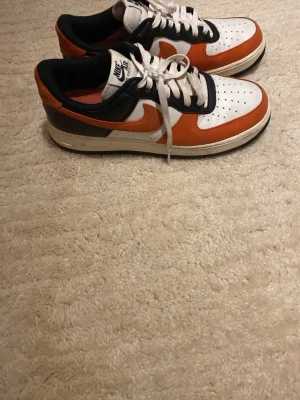 Nike Air Force 1 vit orange och svart - Nike Air Force 1 sneakers i vitt, orange och svart läder. Klassisk låg modell med vit tåbox, orange swoosh och detaljer, samt svart häl och snörning. Tjock vit sula med svart yttersula. Perfekta för dig som vill sticka ut med färg på fötterna.