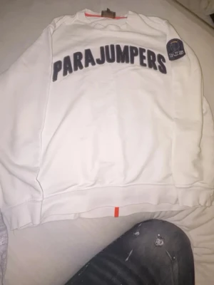 Vit sweatshirt från Parajumpers - Vit sweatshirt från Parajumpers med stor svart logotyp på bröstet och patch på ärmen. Tröjan har rund halsringning och ribbade muddar. Perfekt för en avslappnad och stilren look.
