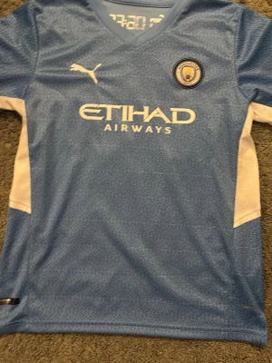 Manchester City matchtröja Puma - Snygg ljusblå Manchester City matchtröja från Puma med klubbmärke och Etihad Airways-tryck på bröstet. Nummer 10 och namnet Grealish på ryggen. V-ringad modell med vita detaljer på sidorna. Perfekt för fotbollsfans!