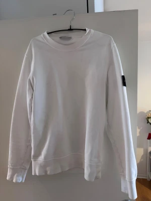 Vit sweatshirt från Stone Island - Vit sweatshirt från Stone Island med klassisk rund hals och lång ärm. Tröjan har en stilren design och ikonisk svart logotyp-patch på vänster ärm. Perfekt för en clean och avslappnad look. Det finns även en QR kod på insidan av tröjan där man kan scanna och få bekräftat att den är äkta. Köpt för ca 3000 kr 