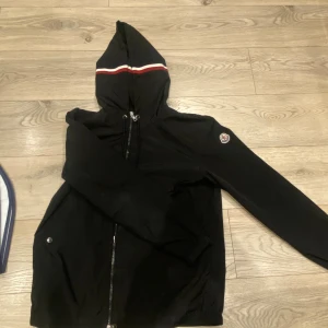 moncler windbreaker  - Svart vindjacka från Moncler med huva och dragkedja framtill. Jackan har Moncler-logga på ärmen och snygga rödvita ränder på huvan. jackan är helt skadefri och nästintill nyskick, har endast använt den ett fåtal gånger i somras men börjar bli för liten för mig. skickar med tags samt påsen och kvitto. hör av er vid frågor!