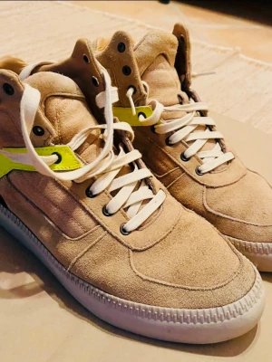 Beige höga sneakers med neon-detalj - Snygga höga sneakers i beige mocka med vita snören och sula. De har en cool neongul detalj på sidan och svart parti vid hälen. Perfekta för dig som vill sticka ut med din stil och gillar streetwear-vibbar.