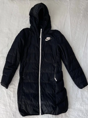 Svart lång dunjacka från Nike - Snygg svart lång dunjacka från Nike med vit dragkedja och logga på bröstet. Jackan har huva, två sidofickor med dragkedja.