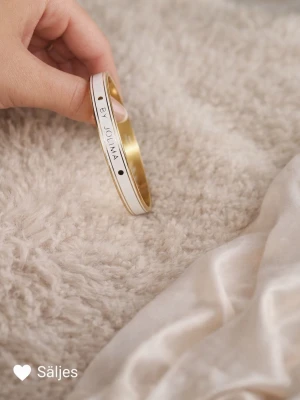 Vitt och guldigt armband By Jolima - Snyggt armband från By Jolima i guldigt metallmaterial med vit emalj på utsidan. Armbandet är stelt och har en stilren design med svarta prickdetaljer och texten BY JOLIMA ingraverad. Perfekt accessoar för att lyfta din outfit. Skriv gärna för mer bilder✨✨✨