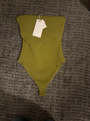 Olivgrön bandeau-body från Zara - Trendig olivgrön bandeau-body från Zara med axelbandslös design och högt skurna ben. Tillverkad i stretchigt material som sitter tight mot kroppen. Perfekt att styla med jeans eller kjol för en snygg look. Aldrig använd.