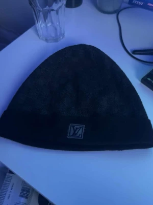 Svart mössa från Louis Vuitton - Snygg svart mössa från Louis Vuitton med diskret LV-logga framtill. Mössan är stickad och har en klassisk form med uppvikt kant. Materialet känns mjukt och bekvämt, perfekt för kyliga dagar. En stilren accessoar som lyfter din outfit.