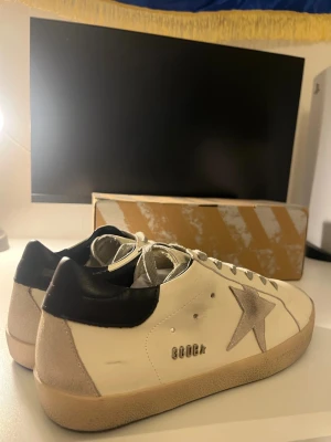 Golden Goose vita sneakers med stjärna - Snygga sneakers från Golden Goose i vitt skinn med grå mockadetaljer och en ikonisk stjärna på sidan. Skorna har svart hälparti, grå snörning och beige platt gummisula. Perfekt för dig som gillar streetstyle och vill ha en unik look.