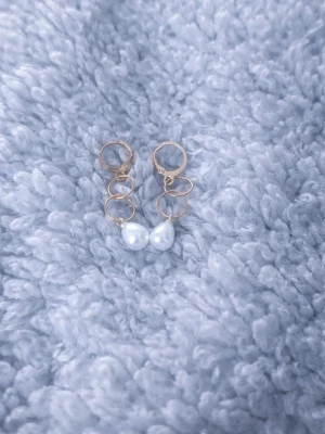 Guldörhängen med vita pärlor - Säljer ett par trendiga örhängen i guldfärgad metall med tre ringar och vita droppformade pärlor längst ner. Perfekta för dig som gillar en elegant men modern stil. Örhängena har en enkel klickstängning och passar till många olika looks.