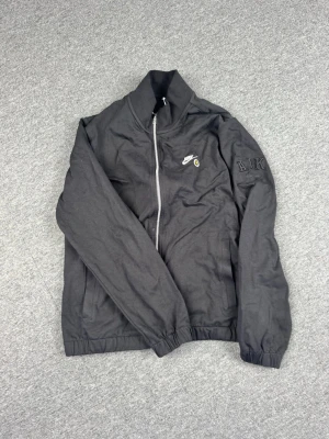 Svart Nike x AIK tröja - Svart tröja från Nike i samarbete med AIK. Jackan har dragkedja framtill, två sidofickor och broderad AIK-logga på ärmen. Nike-logga och liten gul detalj på bröstet. Tillverkad i ett lätt och bekvämt material, perfekt för sport eller chill.