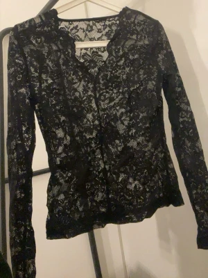 Svart långärmad spetsblus - Snygg svart långärmad topp i genombruten spets med blommigt mönster. Djupare ringning och figurnära passform ger en trendig look. Perfekt att styla lager på lager eller som statement-plagg.