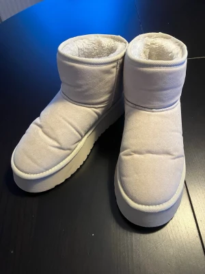 Vita fejk uggs - Säljer ett par vita boots, uggskopior, med fluffigt foder och platt sula. Skorna har en rund tå och är tillverkade i syntetmaterial med mjuk, vadderad utsida. Supervarma och sköna!