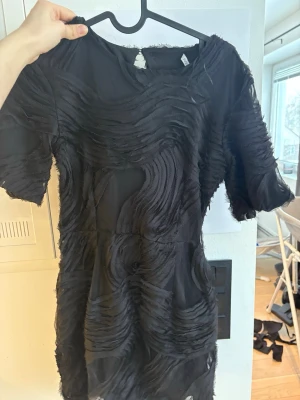 Svart kortklänning med struktur - Svart kortklänning med unik strukturerad yta och korta ärmar. Klänningen har ett vågigt mönster i tyget som ger en cool och modern look. Perfekt för dig som vill sticka ut med en annorlunda stil.