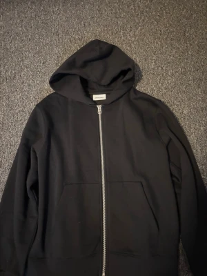 Svart hoodie med dragkedja Jack & Jones - Klassisk svart hoodie från Jack & Jones med dragkedja framtill och två stora fickor. Hoodien har en rymlig huva. Använd 1 gång