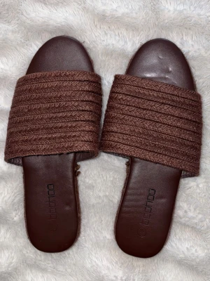 Bruna sandaler/tofflor från Boohoo strl 37 - Säljer ett par bruna sommarskor från Boohoo med bred ribbad rem och platt sula. Perfekta att glida runt i på semester, med en stilren och enkel design.  Storlek 37 och knappt använd