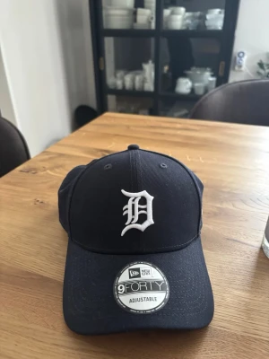 Svart New Era 9FORTY keps - Snygg svart keps från New Era, modell 9FORTY, med justerbar passform och ett vitt broderat D på framsidan. Klassisk böjd skärm och ventilerande hål upptill. Perfekt för en sportig och avslappnad stil. Varan är aldrig använd och är därför som NY, pris u butik 400-500kr mitt pris 250kr! Pris ej hugget i sten vid smidig och snabb affär