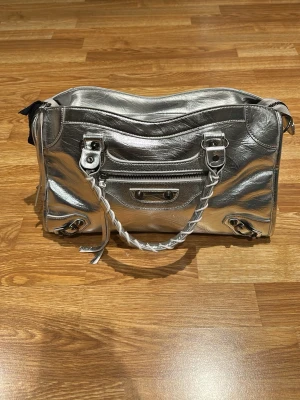 Citybag i silver perfekt skick - Cool handväska i metallic silverfärgat skinn. Väskan har flätade handtag, dragkedja upptill och detaljer i silver. Perfekt för dig som vill sticka ut med en statementväska. Oanvänd.