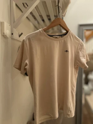 Beige t-shirt med liten gant loga  - En stilren beige t-shirt från gant med rund halsringning och en liten broderad logga på bröstet. Perfekt basic-plagg att matcha med allt. Tillverkad i mjuk bomull och har en klassisk passform med korta ärmar.