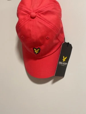 Röd keps från Lyle & Scott - Snygg röd keps från Lyle & Scott med klassisk böjd skärm och justerbar baksida. Kepsen är i bomull och har ventilationshål för extra komfort. Perfekt accessoar för en sportig och trendig look. Helt ny!