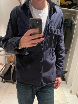 Mörkblå overshirt - Snygg mörkblå overshirt med knäppning framtill och två bröstfickor med lock. Overshirten har en avslappnad passform och är tillverkad i ett mjukt tyg, perfekt att bära över en t-shirt. Enkel och stilren design som funkar till många outfits.