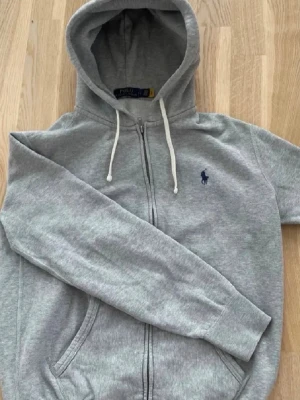Grå zip hoodie Ralph Lauren - Snygg grå hoodie med dragkedja från Polo Ralph Lauren. Klassisk modell med huva och vita dragsnören, två fickor fram och den ikoniska blå loggan broderad på bröstet. Tillverkad i mjuk bomullsblandning som är skön att bära. Lite nopprig men i övrigt perfekt skick! Hör av er angående frågor. 
