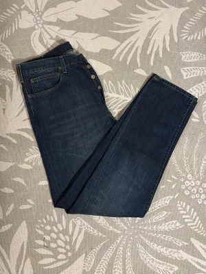 Mörkblå raka jeans från Lee - Snygga mörkblå jeans från Lee med raka ben och klassisk femficksmodell. Jeansen har knäppning med flera synliga knappar och kontrastsömmar. Tillverkade i denim, perfekt för dig som gillar en tidlös och clean look.