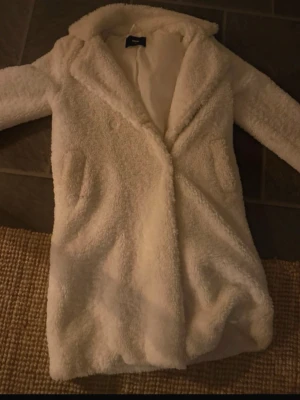 Vit lång teddyjacka med krage - Mysig och fluffig vit teddyjacka med lång passform och bred krage. Jackan har två fickor framtill och stängs med knappar. Perfekt för kyliga dagar när du vill vara både varm och snygg.🩷