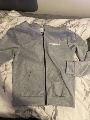 Grå ziptröja från Björn Borg - Snygg grå ziptröja från Björn Borg med svart dragkedja och två fickor framtill. Tröjan har ribbade muddar vid ärmslut och nederkant samt en stilren logga på bröstet. Perfekt till chill eller träning.