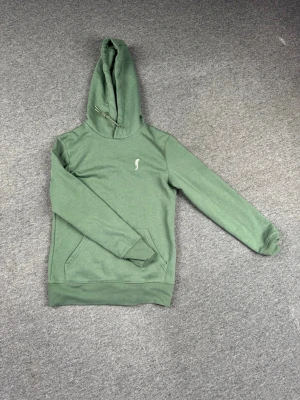 Grön hoodie - RS sports - Snygg grön hoodie med huva och en liten vit logga på bröstet. Hoodien har en klassisk känguruficka framtill. Perfekt för en avslappnad och stilren look. Materialet känns mjukt och bekvämt.