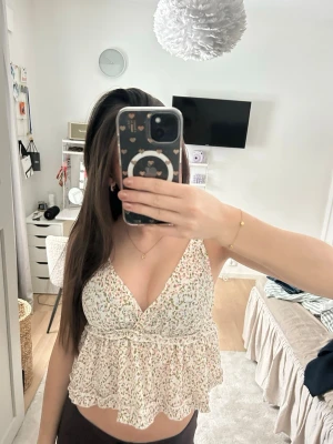 Blommig volangtop med v-ringning - Supersöt blommig topp med volang och v-ringning. Ljus bas med små blommor i grönt, rosa och gult. Ärmlös modell med luftig passform, perfekt för varma dagar. Ger en romantisk och trendig vibe till din outfit. I storlek S men passar en Xs/Xss 