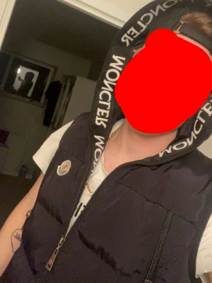 Svart dunväst från Moncler med huva - Snygg svart dunväst från Moncler med stor huva och tydlig logga på bröstet. Västen har två dragkedjeförsedda fickor framtill och Moncler-tryck på huvan. Perfekt för lager-på-lager och streetwear-stil.