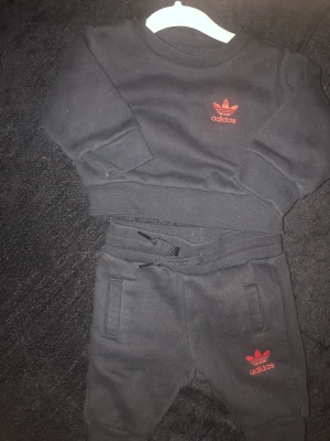 Svart Adidas set med röd logga - Snyggt svart set från Adidas med matchande tröja och byxor. Båda delarna har den klassiska röda Adidas-loggan framtill. Tröjan har rund hals och muddar vid ärmslut, byxorna har dragsko i midjan och fickor. Perfekt för en sportig och avslappnad look.