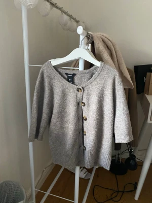 Brun/beige kofta från Lindex med knappar - Mysig beige/brun kofta från Lindex med rund halsringning och stora dekorativa knappar framtill. Koftan har trekvartsärmar och ribbade muddar vid ärmslut och nederkant. 