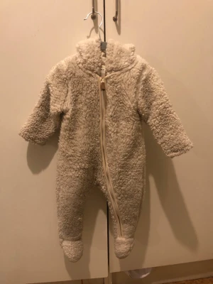 Beige teddyoverall med huva - Supermjuk och fluffig teddyoverall i beige med dragkedja framtill och huva. Overallen har långa ärmar och ben med inbyggda fotdelar, perfekt för kalla dagar. Materialet är teddytyg som ger en mysig känsla.