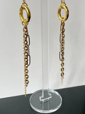 Långa guldiga kedjeörhängen i Rostfritt stål - Säljer ett par långa örhängen i guldton med flera tunna kedjor och ringformad topp. Örhängena har en trendig design med olika kedjelänkar som ger ett coolt och edgy uttryck. Perfekta för dig som vill sticka ut med accessoarerna.