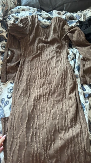 Brun omlottklänning med struktur - Säljer en brun långärmad omlottklänning med snygg struktur i tyget och med bälte i midjan. Klänningen har en unik vågig textur och är i mjukt material som känns skönt mot huden. Perfekt för dig som vill ha något annorlunda och trendigt.