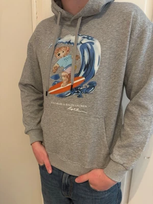 Grå hoodie Polo Bear Ralph Lauren - Grå hoodie från Ralph Lauren med Polo Bear-tryck där björnen surfar på en våg. Klassisk huva med dragsko och magficka framtill. Perfekt för dig som gillar streetstyle och vill sticka ut med en ikonisk print. Han på bilden är 184 cm och väger runt 75 kg. Det är L men skulle säga att den passar dem med M.