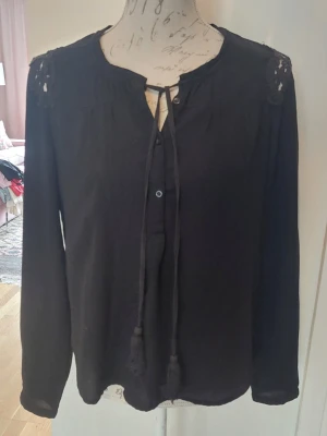 Svart blus med spetsdetaljer - Svart blus med långa ärmar och dekorativa spetsdetaljer på axlar och rygg. Blusen har knytband vid halsen och en lös, flowy passform. Perfekt för dig som gillar en stilren look med lite extra detaljer.