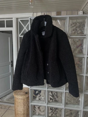 Svart teddyjacka med hög krage - Mysig svart teddyjacka med rak passform. Jackan har en fluffig, fårullsliknande yta och stängs med knappar framtill. Perfekt för kyliga dagar när du vill vara både varm och snygg. Så fint skick 