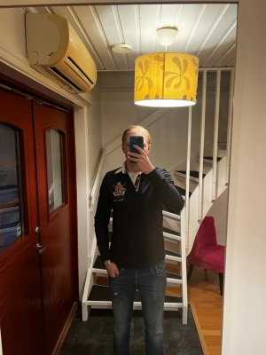 Svart pikétröja  - Svart pikétröja med vit krage och broderad logga på bröstet. Tröjan har knappslå framtill och detaljer i rött och guld. Perfekt för en stilren och sportig look.