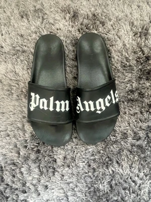 Palm Angels svarta slides - Svarta slides från Palm Angels med bred rem över foten och vit logotyp i gotisk stil. Tillverkade i syntetmaterial med platt sula och öppen tå. Perfekta för chill dagar eller när du vill ha en streetwear-vibe.