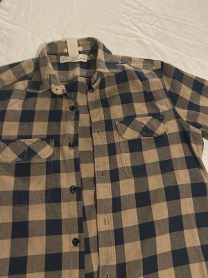 Rutig skjorta från H&M L.O.G.G. S - Säljer en klassisk rutig skjorta från H&M L.O.G.G. i beige och mörkblå. Skjortan har två bröstfickor med lock, svarta knappar och normal passform. Tillverkad i mjuk bomull, perfekt för en avslappnad stil.