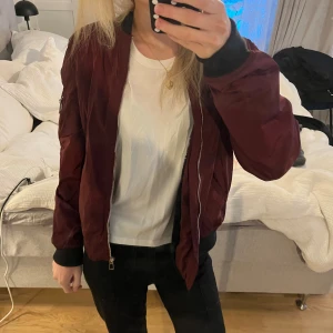 Vinröd bomberjacka från George - Säljer en vinröd bomberjacka från George med klassisk siluett. Jackan har svarta muddar vid ärmslut och midja, dragkedja framtill och en ficka med dragkedja på ena ärmen. Materialet är polyester och jackan är långärmad. Jackan är fodrad med päls. Står ingen storlek men skulle säga S