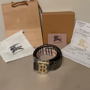 Vändbart bälte (beige/svart) med guldlogga - Exklusivt vändbart bälte med två sidor: beige mönstrad sida samt svart för en mer diskret look. Guldlogga som ger en elegant och lyxig touch. Ett tidlöst accessoarval som lyfter vilken outfit som helst.