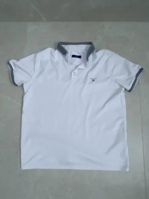 Vit piké från GANT med grå krage - Säljer en stilren vit pikétröja från GANT i storlek M. Pikén har korta ärmar och en gråmelerad krage med matchande detaljer vid ärmsluten. Klassisk GANT-logga broderad på bröstet. Perfekt för en fräsch och enkel look.