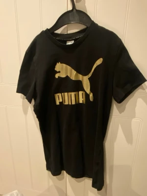 Svart t-shirt med guldtryck från Puma - Svart t-shirt från Puma med stort guldigt logotryck på bröstet. Klassisk rund halsringning och korta ärmar. Tillverkad i mjuk bomull för skön känsla och snygg passform. Perfekt för dig som gillar sportig och stilren look.