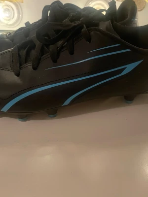 Svarta fotbollsskor från Puma - Säljer ett par svarta fotbollsskor från Puma med blå detaljer och snörning. Skorna har rund tå och platt sula med dobbar för bra grepp på planen. Materialet är syntet och designen är sportig med stilrena linjer längs sidorna.